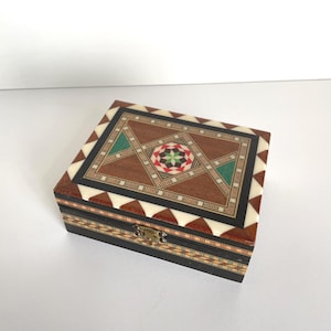 May include: A small, square wooden box with intricate inlaid geometric patterns. The lid features a central starburst design in red, green, and brown, surrounded by a border of white triangles and black lines. The box is closed with a small metal clasp.