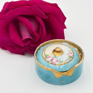 May include: A round, light blue porcelain trinket box with a gold-trimmed lid featuring a hand-painted pink rose floral pattern and a small gold knob. The base of the box also has gold trim and a gold bow-shaped detail on the front side.