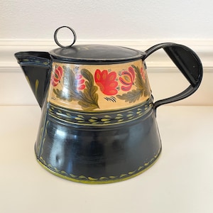 May include: Black and gold metal teapot with a floral design. The teapot features a black handle, spout, and base, with a cream-colored band adorned with red flowers and green leaves. A small ring handle sits atop the lid.