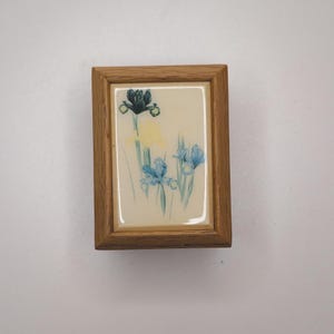 May include: A small, rectangular wooden frame with a floral print. The print features blue, yellow, and green irises on a cream-colored background. The frame has a light brown, natural wood finish.