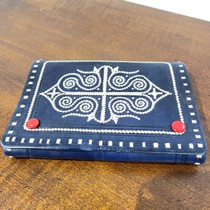 May include: A navy blue leather wallet with white embroidered geometric designs and red button closures. The wallet has a rectangular shape with a detailed pattern on the front and white stitching around the edges.