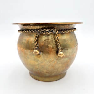 May include: A round, hammered brass pot featuring a wide, flat rim and a decorative rope-like band encircling the upper section. The rope detail includes two hanging ends that terminate in small, smooth, spherical brass finials on the front.