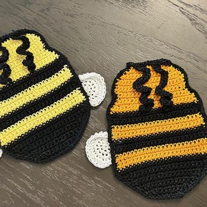 May include: Two handmade crochet bee-shaped coasters. Each coaster features a black base with yellow stripes, a yellow top, and white wings. The coasters are designed to resemble bees and are suitable for home decor.