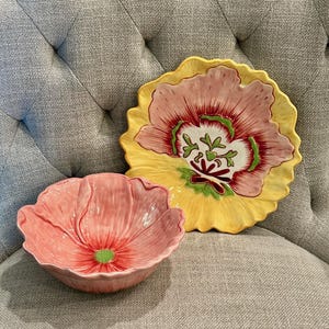 May include: A ceramic bowl and a matching plate shaped like flowers. The bowl is pink with a green center. The plate features a large pink flower design with yellow petals, green accents, and dark red details, both with scalloped edges.