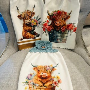 Könnte beinhalten: Drei weiße Hängeschirrtücher mit gehäkelten Oberteilen in Creme, Dunkelrot und Blaugrün. Jedes Tuch zeigt eine gedruckte Illustration eines Hochlandrindes: eines mit Blumen, eines in einem Metalleimer mit Rosen und eines in einem Korb mit Ostereiern.