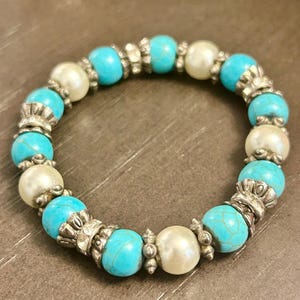 May include: A beaded bracelet featuring alternating turquoise and pearl-like beads. The turquoise beads are round and have a textured appearance. The bracelet is strung with silver-colored metal spacers.