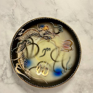 May include: A round decorative plate featuring a central cream-colored field with a black gradient border. A detailed dragon illustration in gold, white, and blue wraps around the center, accented by stylized gold flame motifs and black line art.