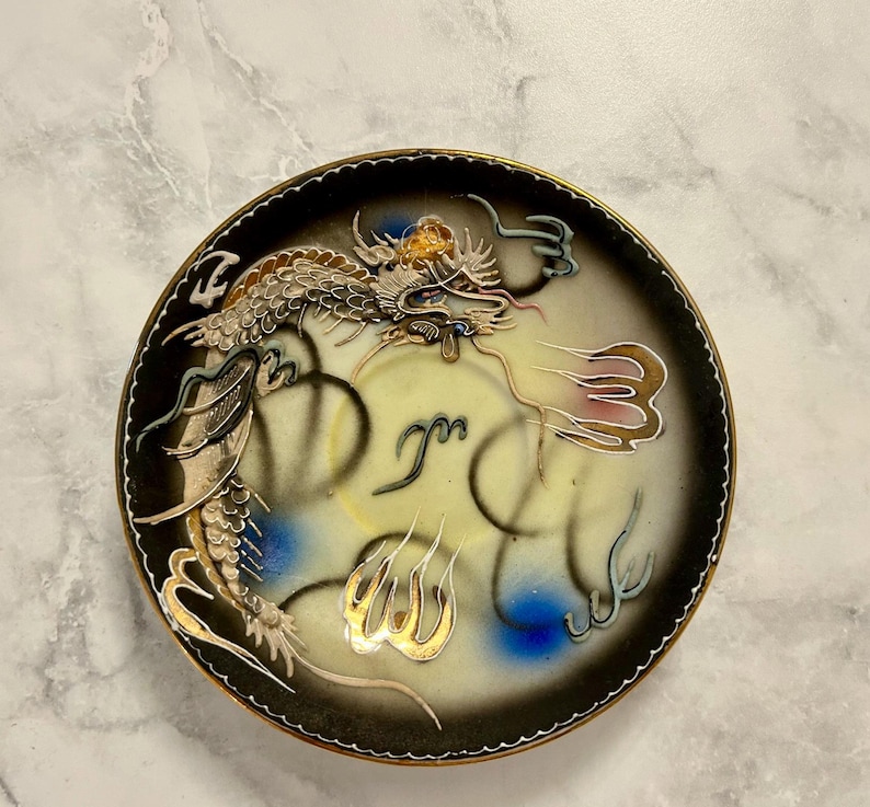 May include: A round decorative plate featuring a central cream-colored field with a black gradient border. A detailed dragon illustration in gold, white, and blue wraps around the center, accented by stylized gold flame motifs and black line art.