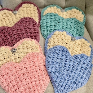 May include: A collection of handmade crochet heart-shaped items in various colors, including pink, purple, blue, and cream. Each heart features a textured stitch pattern and a decorative embellishment. These items are likely decorative or functional, such as coasters or trivets.