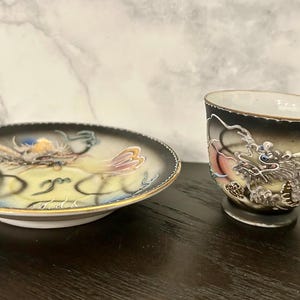 May include: A porcelain teacup and matching saucer set featuring a raised, textured dragon motif in silver, gold, and blue tones against a black and cream gradient background, finished with a delicate gold rim on both the cup and the saucer edges.