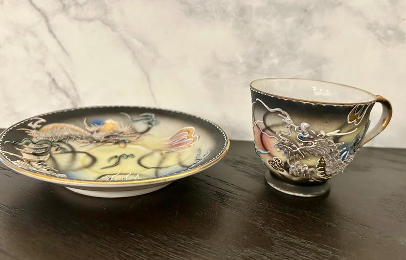 May include: A porcelain teacup and matching saucer set featuring a raised, textured dragon motif in silver, gold, and blue tones against a black and cream gradient background, finished with a delicate gold rim on both the cup and the saucer edges.