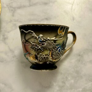May include: A ceramic teacup featuring a raised, three-dimensional dragon motif in silver, gold, and blue tones against a dark, gradient background. The cup has a gold-trimmed handle and a scalloped rim, showcasing intricate relief detailing.