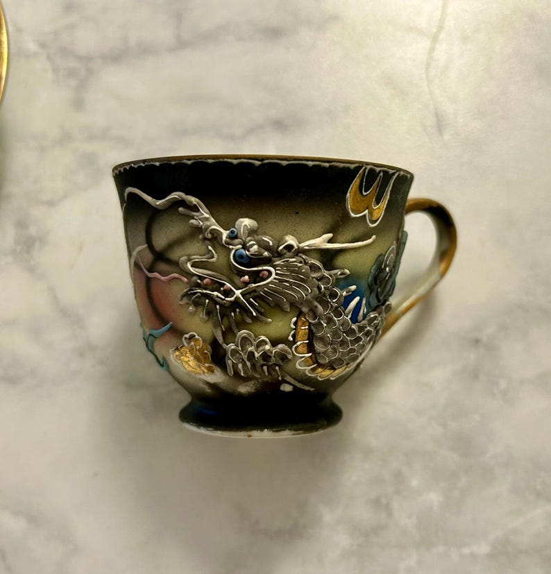 May include: A ceramic teacup featuring a raised, three-dimensional dragon motif in silver, gold, and blue tones against a dark, gradient background. The cup has a gold-trimmed handle and a scalloped rim, showcasing intricate relief detailing.