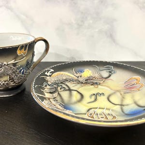 May include: A ceramic teacup and matching saucer set featuring a raised, textured dragon motif. The design incorporates a gradient of black, cream, and blue tones with gold-colored rim accents. The dragon appears in relief on both pieces.