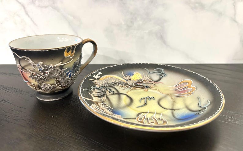 May include: A ceramic teacup and matching saucer set featuring a raised, textured dragon motif. The design incorporates a gradient of black, cream, and blue tones with gold-colored rim accents. The dragon appears in relief on both pieces.