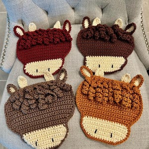 May include: Four handmade crochet coasters in the shape of a cow's head. Each coaster features a different shade of brown, with cream-colored faces and ears. The coasters are arranged on a gray upholstered chair.