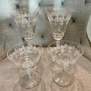 May include: Four clear glass stemmed cocktail glasses with etched floral designs. Two glasses have a flared shape, and two have a shallow, rounded bowl. The glasses are arranged on a gray upholstered chair.