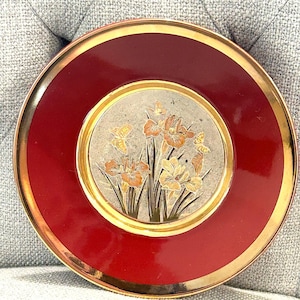 May include: A round decorative plate featuring a deep red border with a gold rim. The center is a textured, light-colored circular panel decorated with an illustration of orange and yellow iris flowers and small butterflies in a gold-toned finish.