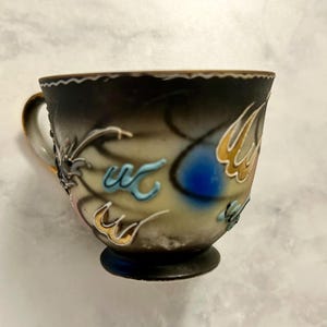 May include: A ceramic teacup featuring a gradient finish that transitions from a dark charcoal rim to a cream-colored base. The exterior is decorated with raised, abstract relief patterns in shades of blue, gold, and white, with a gold-trimmed handle.