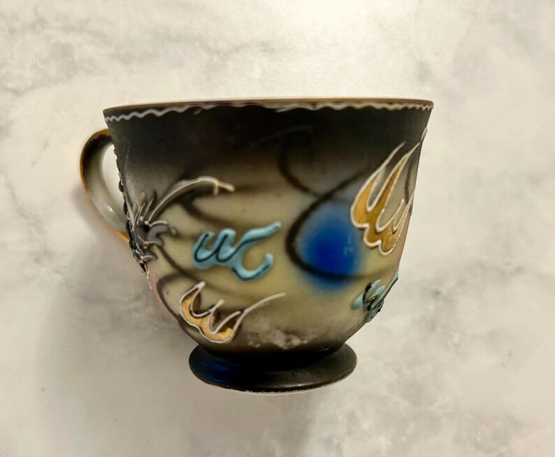 May include: A ceramic teacup featuring a gradient finish that transitions from a dark charcoal rim to a cream-colored base. The exterior is decorated with raised, abstract relief patterns in shades of blue, gold, and white, with a gold-trimmed handle.