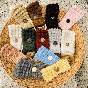 Könnte beinhalten: Eine Sammlung von zwölf rechteckigen Häkel-Handtuchhaltern in verschiedenen Farben wie Beige, Braun, Schwarz, Rosa, Weiß, Grün, Rost, Grau, Blau und Gelb. Jeder Halter verfügt über ein strukturiertes Noppenmuster und einen Zierknopf.