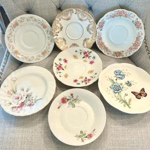 May include: A collection of six round ceramic saucers and plates featuring various designs. One saucer has gold filigree, others display floral patterns in pink, yellow, and blue, and one plate features a butterfly and blue poppy flowers.