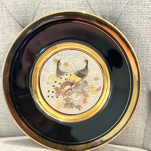 May include: A round decorative plate featuring a glossy black rim with a gold-toned inner border. The center medallion displays a detailed illustration of two peacocks surrounded by various flowers in shades of pink, yellow, and white on a textured background.