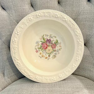 May include: Cream-colored ceramic serving bowl with a floral design in the center. The bowl has a raised, decorative border with a grape vine pattern. The floral arrangement features pink, purple, and yellow flowers.
