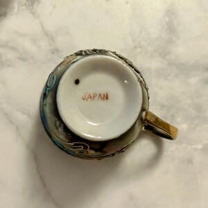 May include: A ceramic teacup viewed from the bottom, featuring a white base with the red text JAPAN printed in the center and a small dark brown dot. The exterior of the cup is dark with metallic gold, blue, and white decorative relief details.