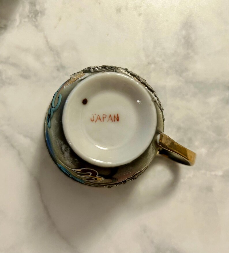 May include: A ceramic teacup viewed from the bottom, featuring a white base with the red text JAPAN printed in the center and a small dark brown dot. The exterior of the cup is dark with metallic gold, blue, and white decorative relief details.