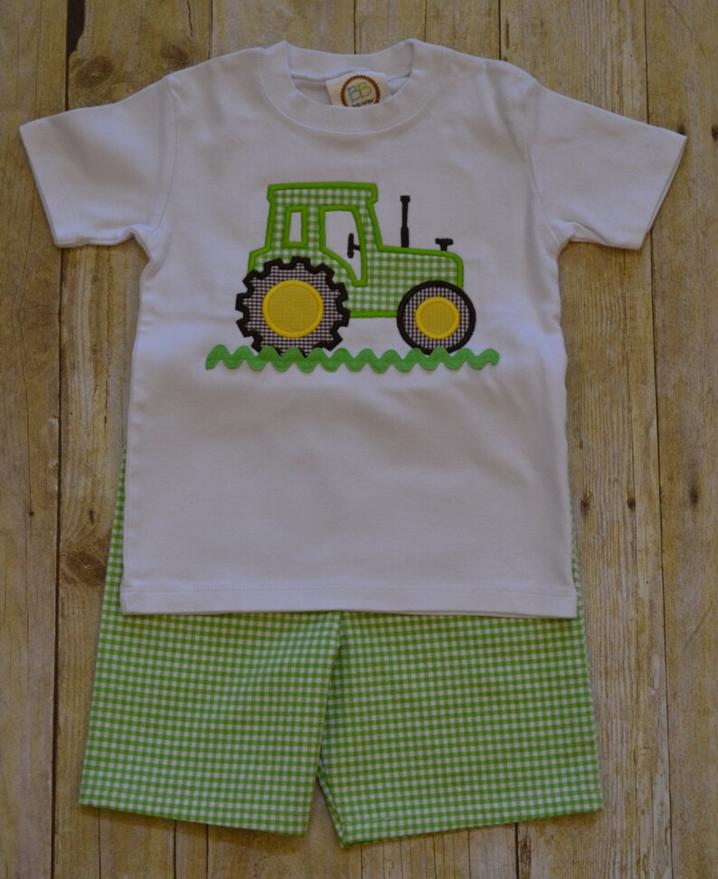 K&ouml;nnte beinhalten: Ein wei&szlig;es T-Shirt mit einem gestickten Traktor-Design in Gr&uuml;n und Schwarz. Das Shirt wird mit gr&uuml;n-wei&szlig; karierten Shorts kombiniert.