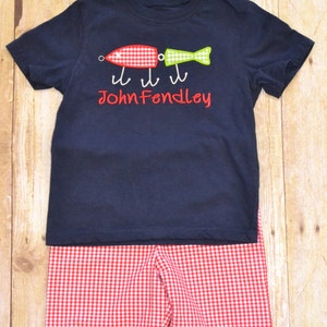 K&ouml;nnte beinhalten: Ein marineblaues T-Shirt mit einem roten und gr&uuml;nen Fischk&ouml;der-Design und dem Namen "John Fendley", der auf der Vorderseite gestickt ist. Das Shirt wird mit rot-wei&szlig; karierten Shorts kombiniert.