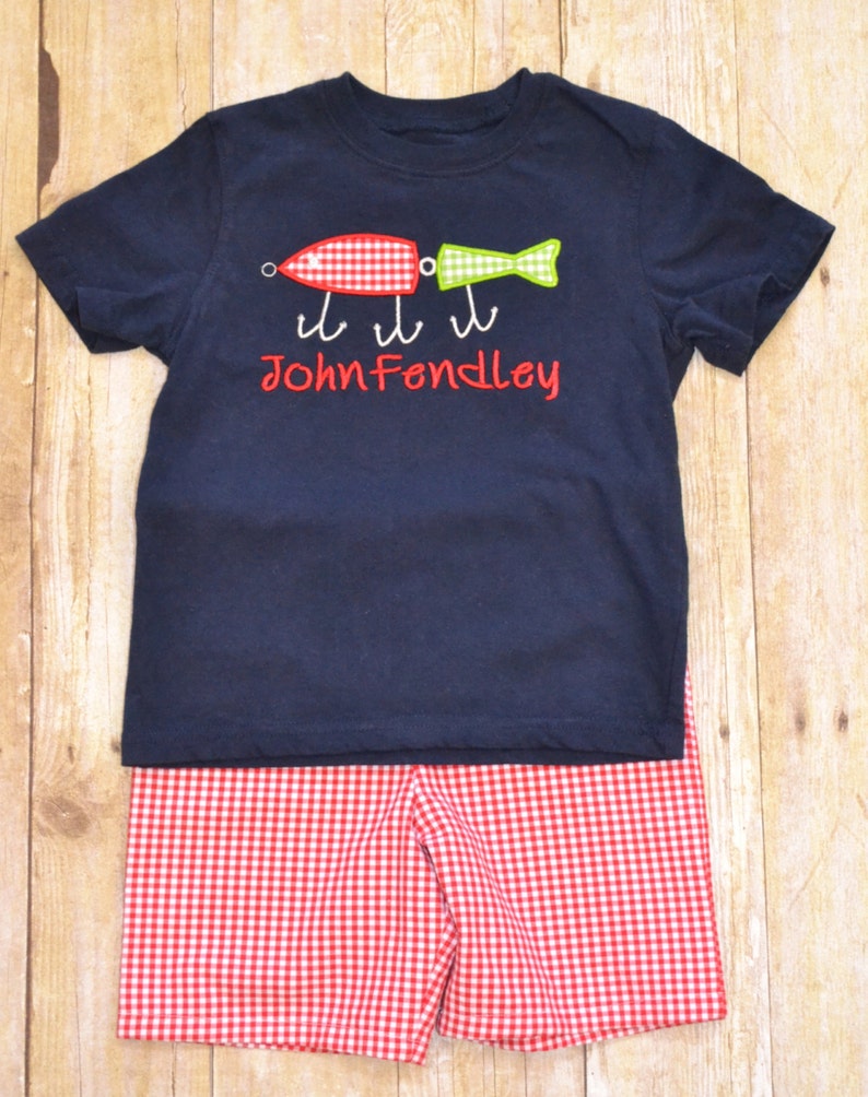 K&ouml;nnte beinhalten: Ein marineblaues T-Shirt mit einem roten und gr&uuml;nen Fischk&ouml;der-Design und dem Namen "John Fendley", der auf der Vorderseite gestickt ist. Das Shirt wird mit rot-wei&szlig; karierten Shorts kombiniert.