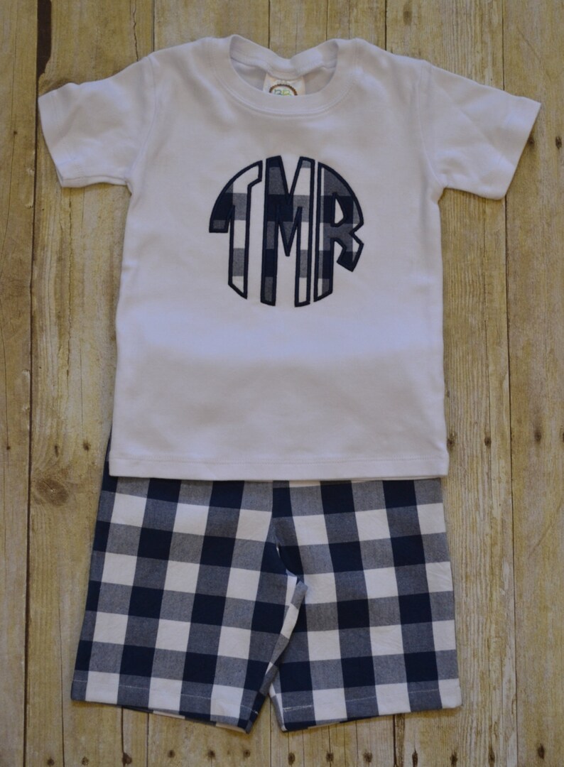 K&ouml;nnte beinhalten: Ein wei&szlig;es T-Shirt mit kurzen &Auml;rmeln und einem marineblauen und wei&szlig;en karierten Monogramm auf der Vorderseite. Das Shirt wird mit marineblauen und wei&szlig;en karierten Shorts kombiniert.