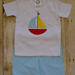 K&ouml;nnte beinhalten: Wei&szlig;es T-Shirt mit kurzen &Auml;rmeln und einem gestickten Segelboot-Motiv in Rot, Gelb und Blau. Das Shirt wird mit blau-wei&szlig; karierten Shorts kombiniert.