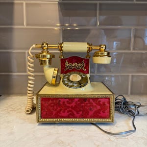 May include: A vintage-style rotary telephone featuring a cream-coloured base and handset with gold-tone metal accents. The front panel and upper section are decorated with a deep red velvet-like fabric featuring an ornate, embossed floral pattern.