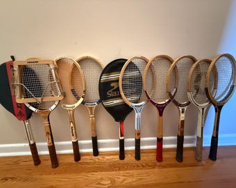May include: A collection of nine vintage wooden tennis rackets arranged in a row. Some rackets feature protective covers with brand names like Dunlop and Slazenger, while others show exposed wooden frames, nylon strings, and various grip colours.