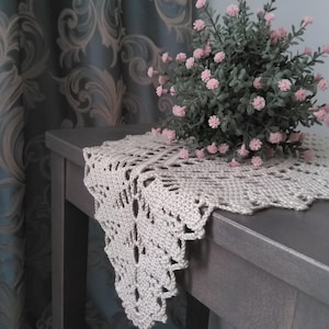 Puede incluir: Un tapete beige tejido a ganchillo con borde festoneado descansa sobre una mesa gris. Un pequeño arbusto con hojas verdes y flores rosas se encuentra encima del tapete. El fondo presenta una cortina estampada.