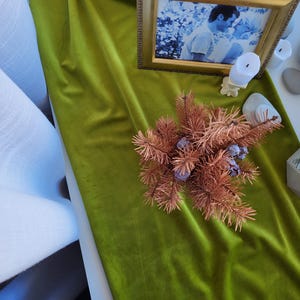May include: A decorative display featuring a gold-framed photograph, a white candle, and a dried floral arrangement. The items are arranged on a vibrant green velvet tablecloth. Small white and gray stones are also present.