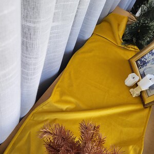 Puede incluir: Un camino de mesa de terciopelo amarillo con una rama de pino marrón en la parte superior. El camino de mesa está drapeado sobre una superficie de madera y una cortina blanca transparente está en el fondo.