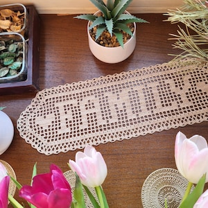 Könnte beinhalten: Ein beigefarbener gehäkelter Tischläufer mit einem gewellten Rand. Der Läufer hat das Wort "Willkommen" in das Design gehäkelt. Der Läufer liegt auf einem Holztisch mit einem Strauß rosa und roter Tulpen und einer Sukkulente im Topf.