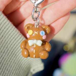 May include: A brown teddy bear keychain with a white bow tie, stitched details, and colorful accents. The bear has a yellow and blue eye, pink cheeks, and a silver clasp.