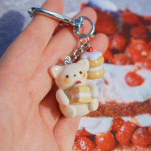 May include: A cream-colored bear keychain with a small cake in its paws and a cupcake charm. The bear has brown freckles and a red dot on its mouth. The keychain is attached to a silver clasp.