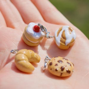 May include: A collection of miniature food charms, including a layered pastry with white frosting and a red cherry, a cinnamon roll with white icing, a croissant, and a chocolate chip cookie. Each charm has a silver-colored loop.