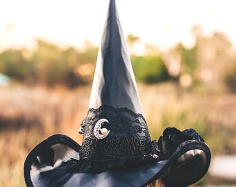 May include: A black witch hat with a wide brim and a tall pointed crown. The hat is decorated with black lace and a silver crescent moon charm.