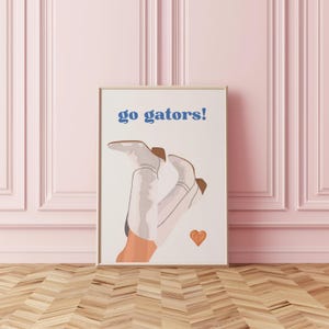 Peut inclure: Impression d'art encadr&eacute;e avec le texte "go gators!" en bleu. L'&oelig;uvre pr&eacute;sente une paire de bottes de cowboy blanches avec des accents marron et un petit c&oelig;ur orange. Les bottes sont positionn&eacute;es avec les talons en l'air. Le fond est blanc.