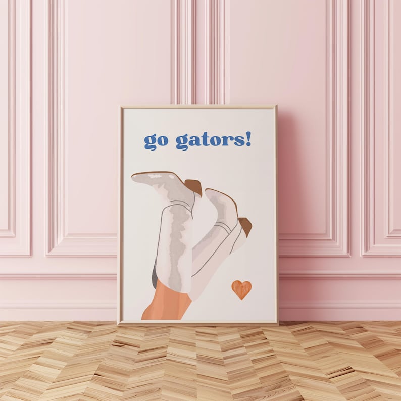 Peut inclure: Impression d'art encadr&eacute;e avec le texte "go gators!" en bleu. L'&oelig;uvre pr&eacute;sente une paire de bottes de cowboy blanches avec des accents marron et un petit c&oelig;ur orange. Les bottes sont positionn&eacute;es avec les talons en l'air. Le fond est blanc.
