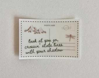 May include: A rectangular postcard with a mountain and tree illustration, a dragonfly, and the words "look at you go, crossin' state lines with your shadow." The word "POSTCARD" is printed at the top.