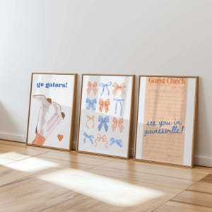 Peut inclure: Trois impressions d'art encadr&eacute;es avec des cadres dor&eacute;s. L'impression de gauche dit "go gators!" au-dessus d'un dessin de botte. L'impression du milieu pr&eacute;sente des n&oelig;uds aquarelles bleus et oranges. L'impression de droite est un re&ccedil;u avec le texte "see you in gainesville!"