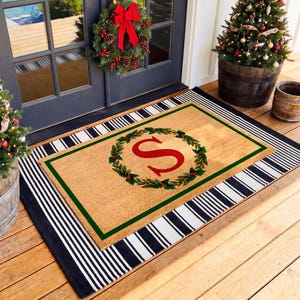 May include: A rectangular tan coir doormat layered over a black and white striped rug. The doormat features a green border, a central green wreath with red berries, and a large red letter S in the center, creating a festive holiday appearance.